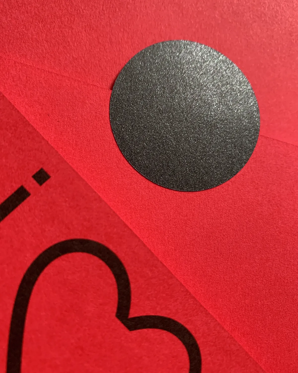 Close-up of a red envelope sealed with a round black sticker, with a card showing a black heart outline below.